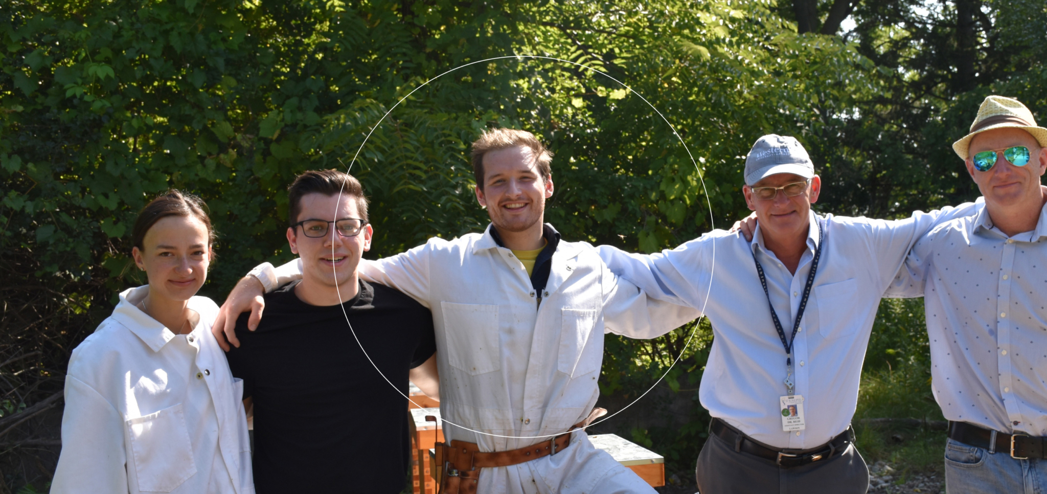 Group of five people standing outdoors with arms around each other, smiling in daylight.