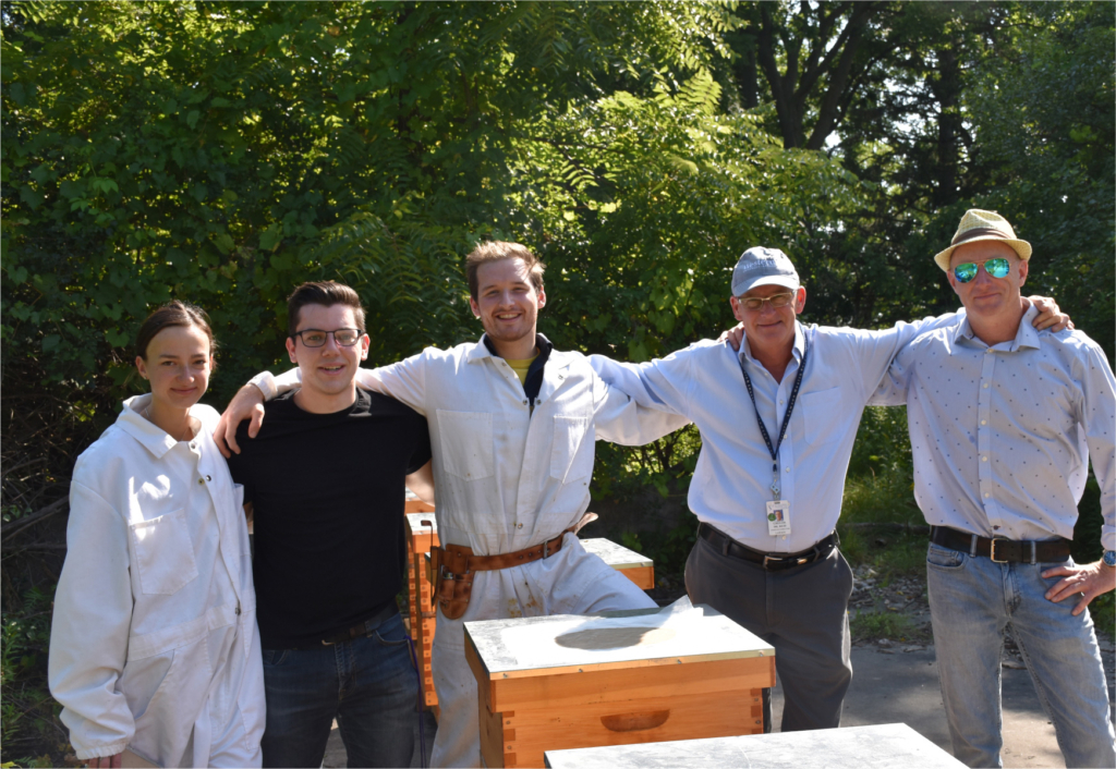 Group of five people standing outdoors with beekeeping equipment and greenery behind them