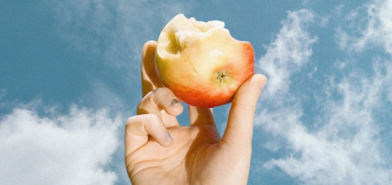 Hand holding a partially eaten apple against a blue sky with clouds.