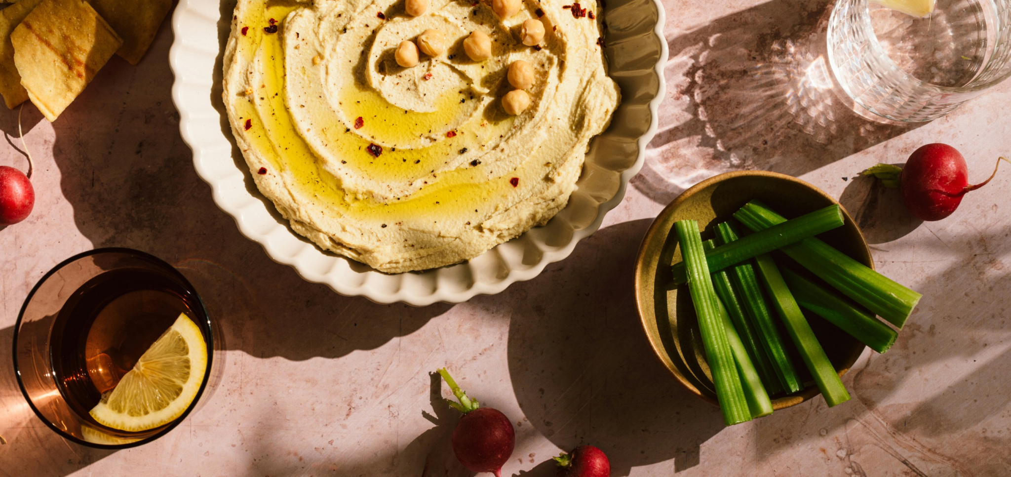 Bowl of hummus with olive oil and chickpeas, celery sticks, radishes, lemon water, and pita chips.