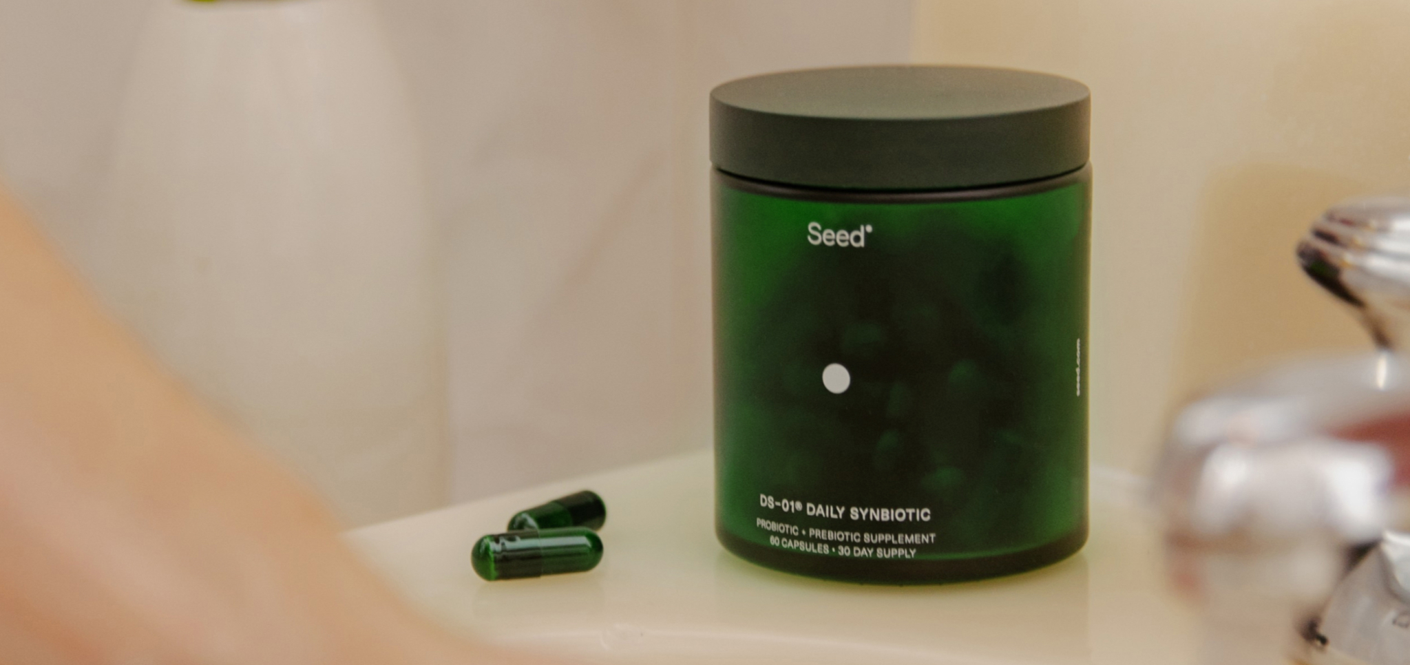 Green container of daily synbiotic supplement with two capsules on a bathroom sink.