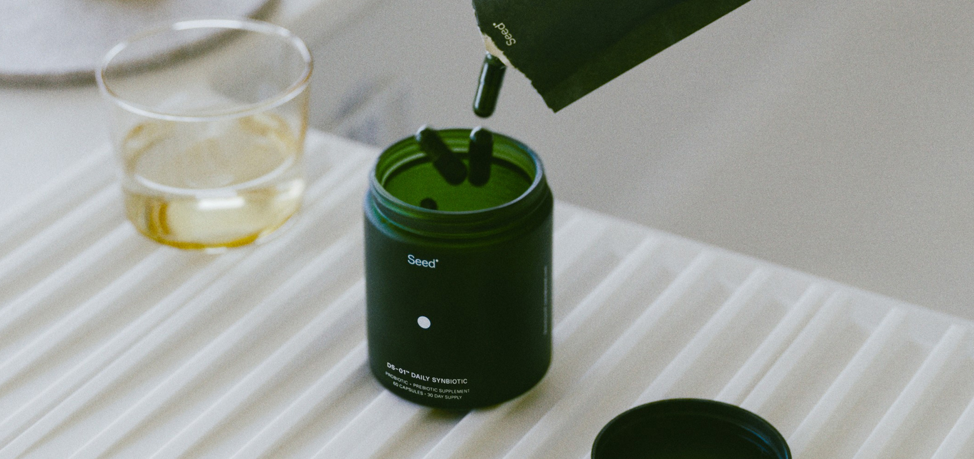Green supplement bottle with capsules being poured in, next to a glass of liquid.