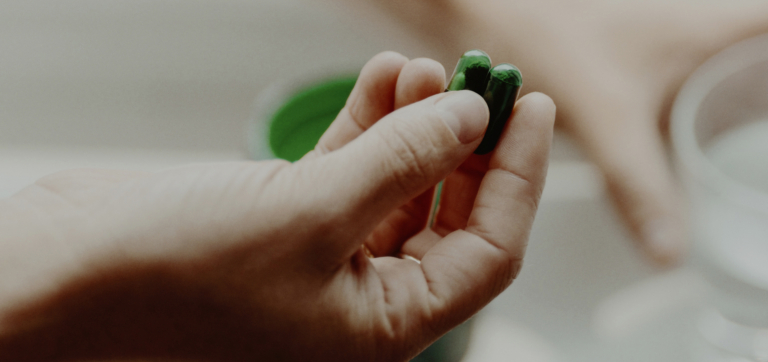 Hand holding two green capsules with a glass of water in the background.