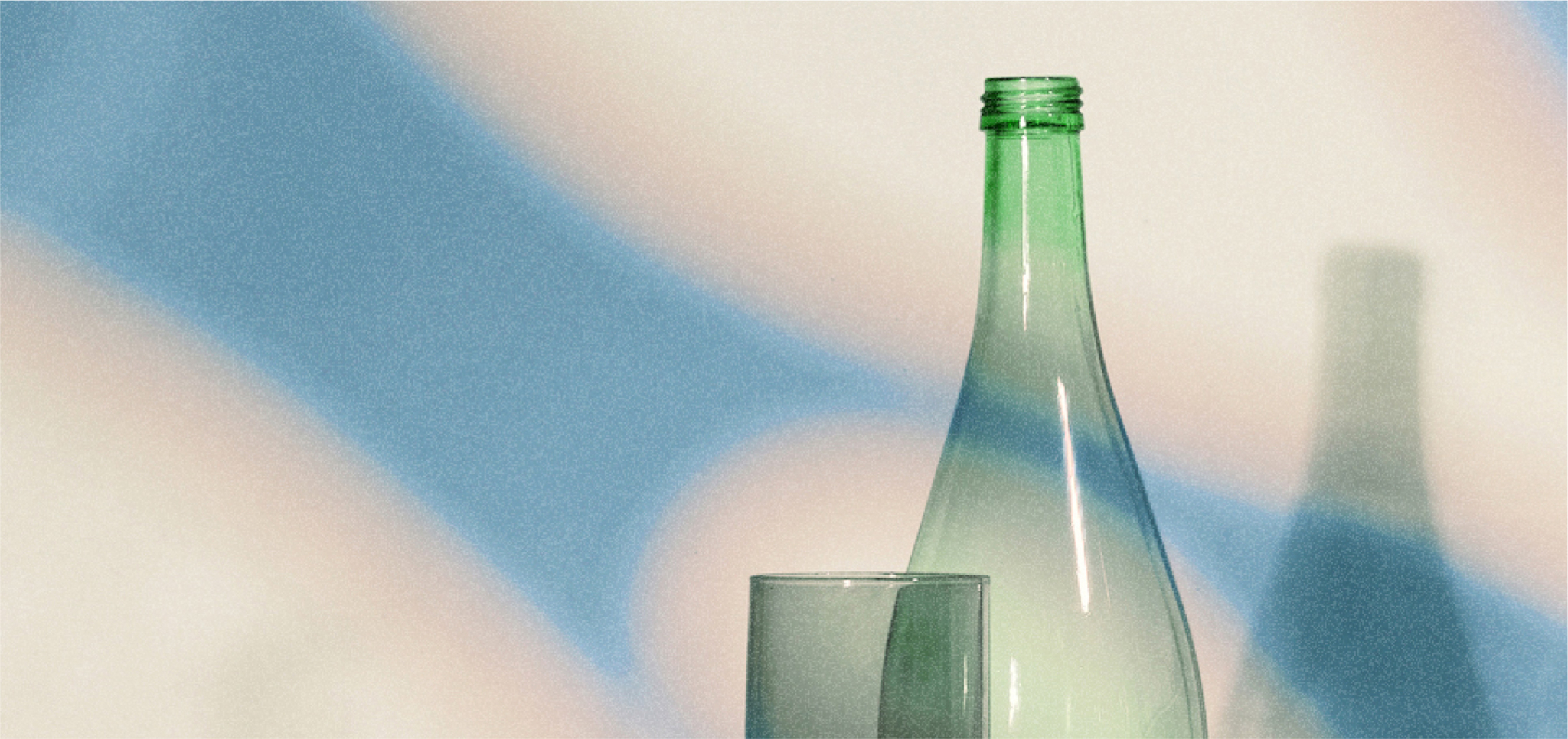 Green glass bottle and glass with blue and beige light patterns in background