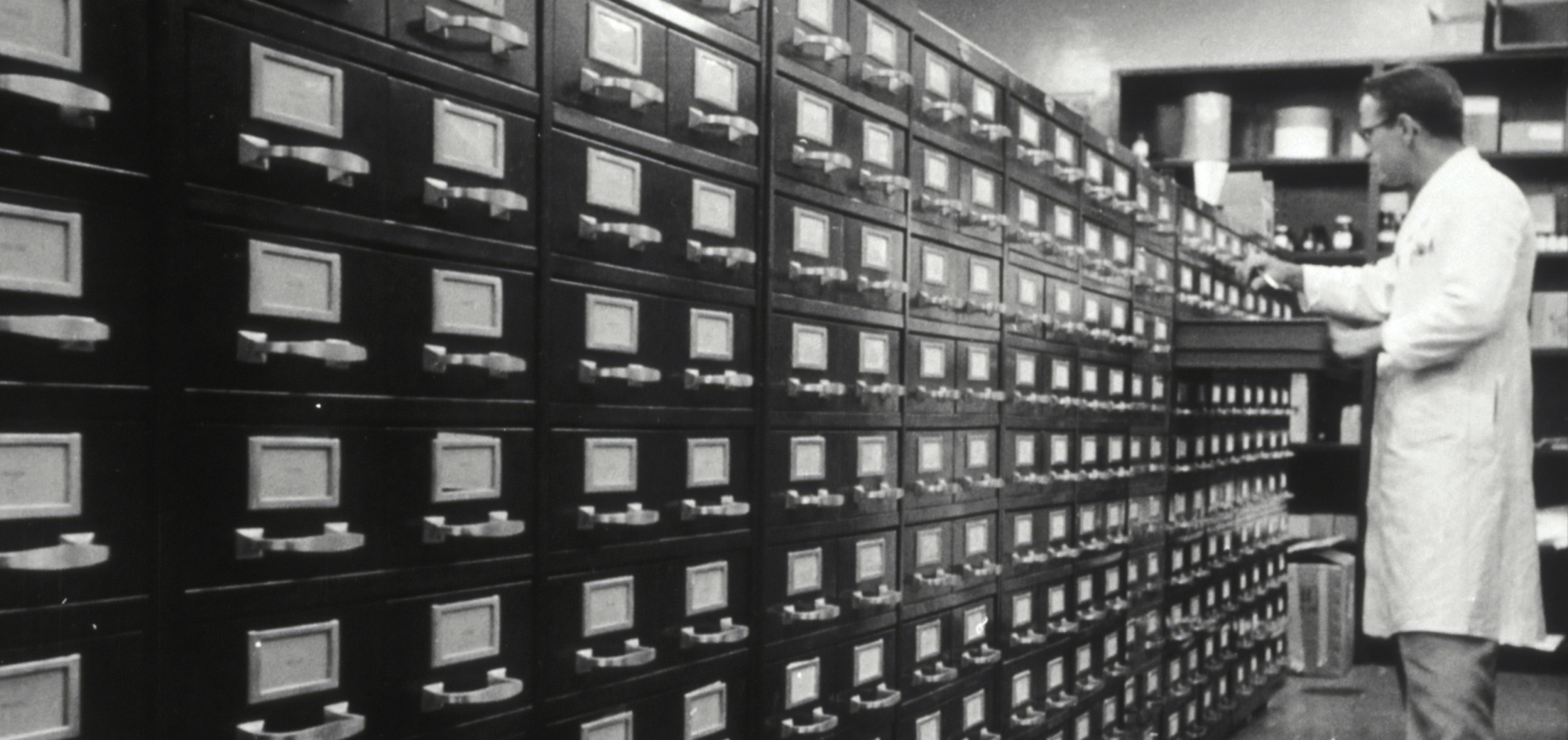 Man in lab coat accessing drawers in a large filing cabinet in a storage room.