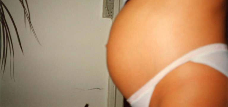 Close-up of a pregnant belly with white underwear against a plain wall background.