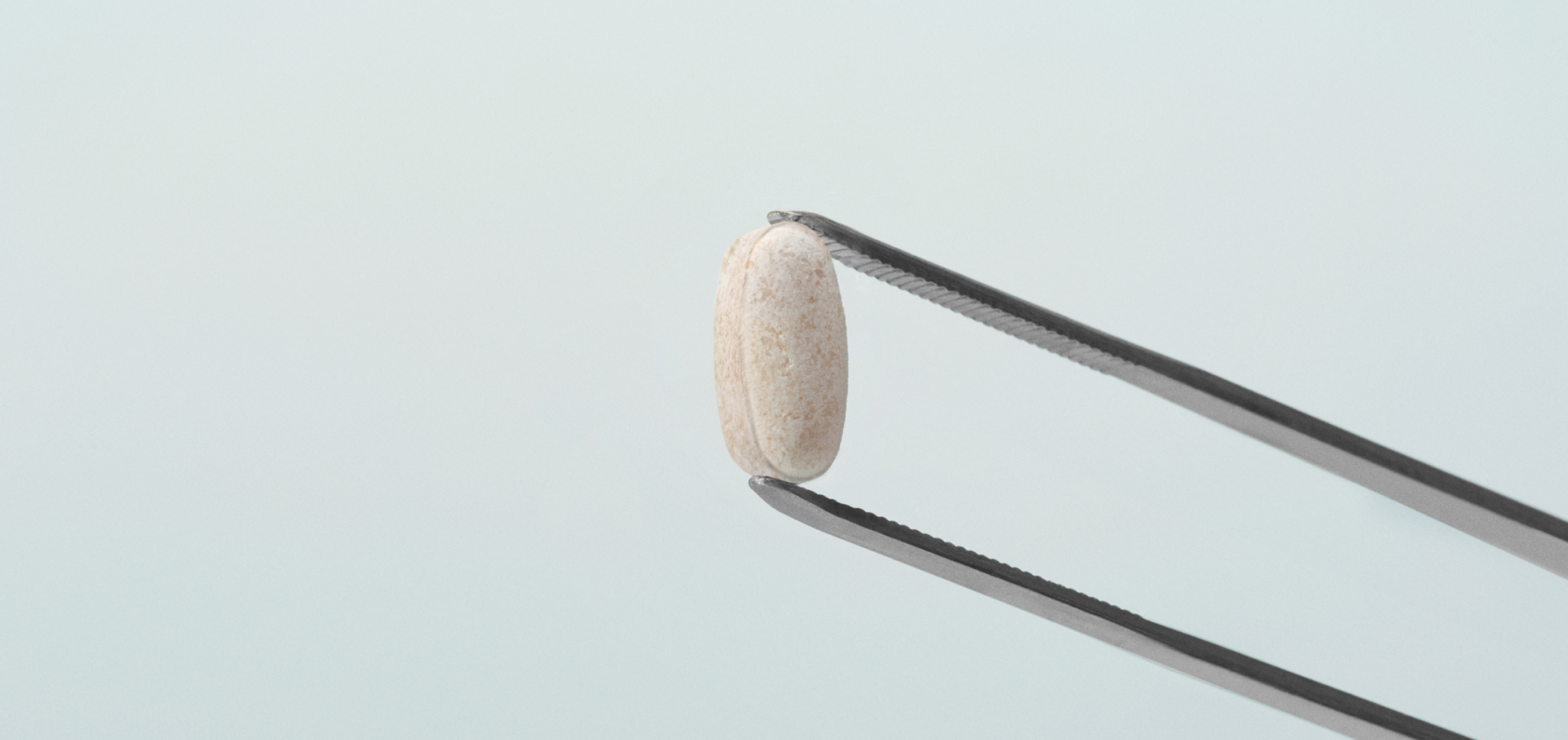 Metal tweezers holding a single beige oval pill against a light background