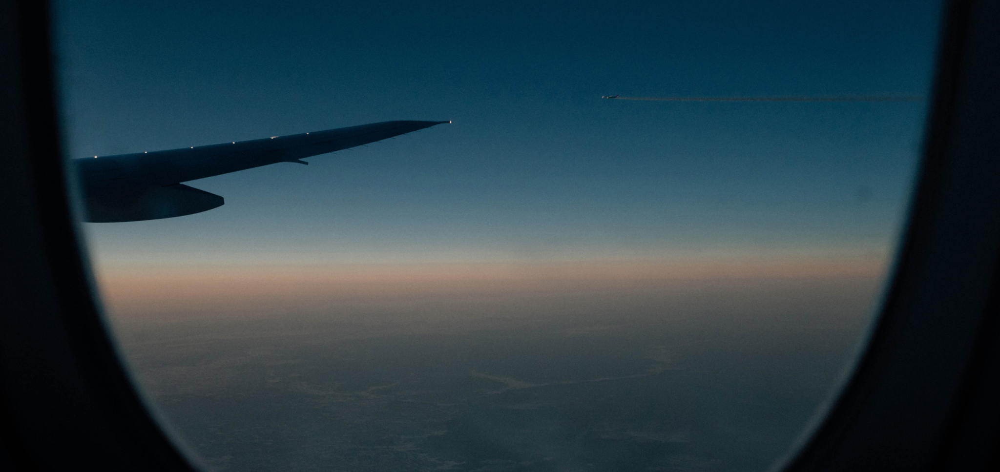 Airplane wing and distant plane flying at sunset viewed through window.