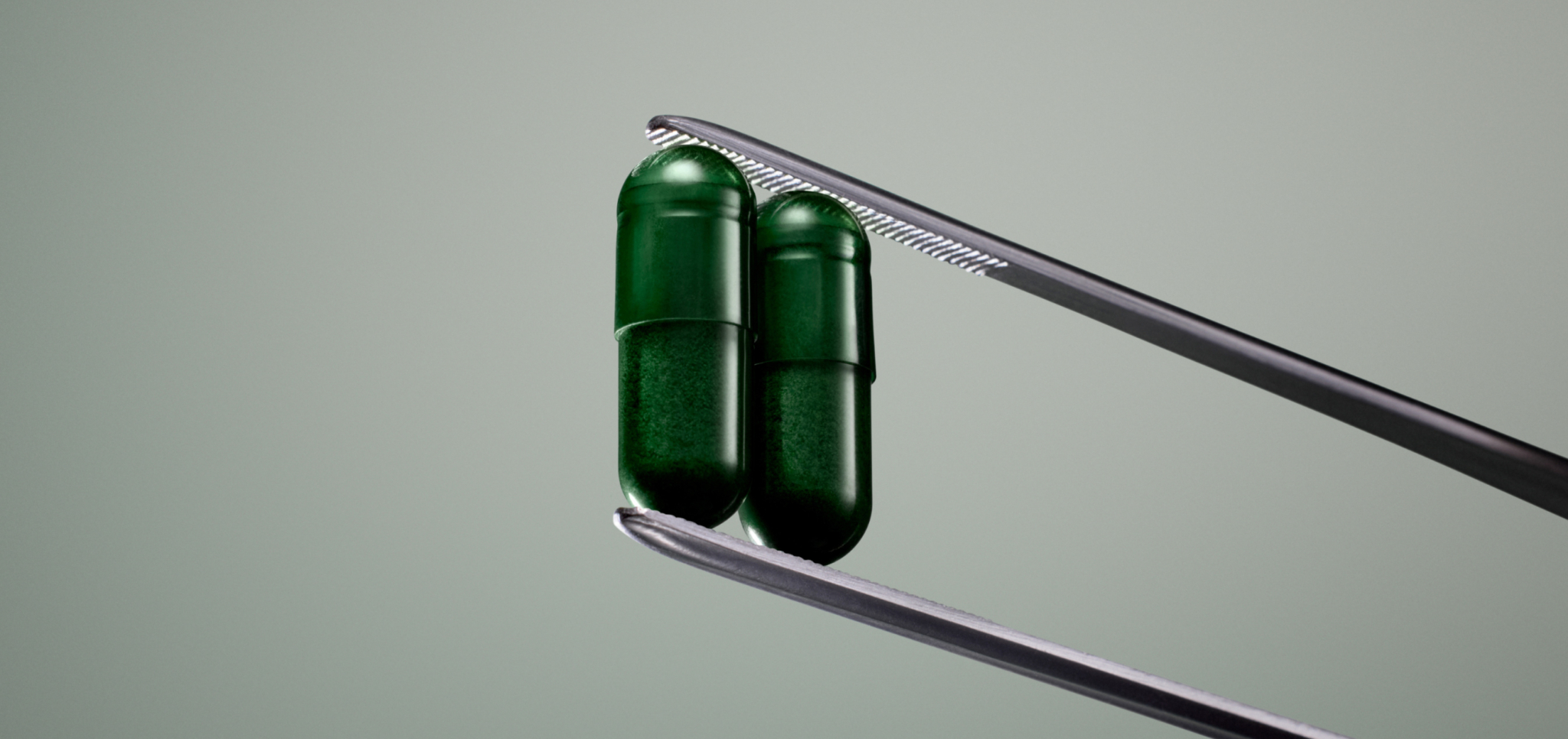 Two green capsules held by metal tweezers against a plain background.