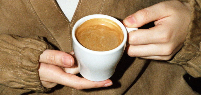 Hands holding a white cup filled with espresso coffee against a brown jacket background