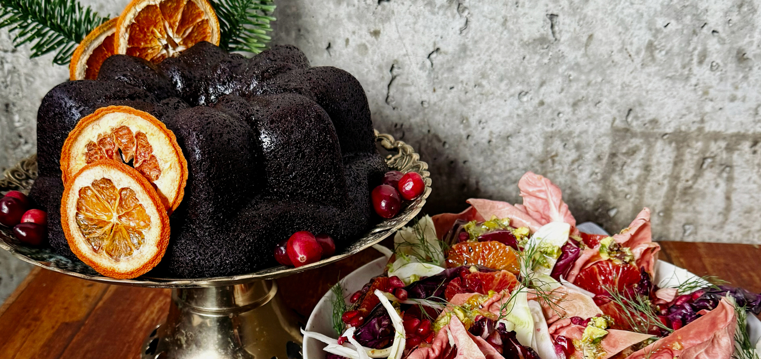 Dark bundt cake garnished with dried orange slices and cranberries next to a colorful salad.