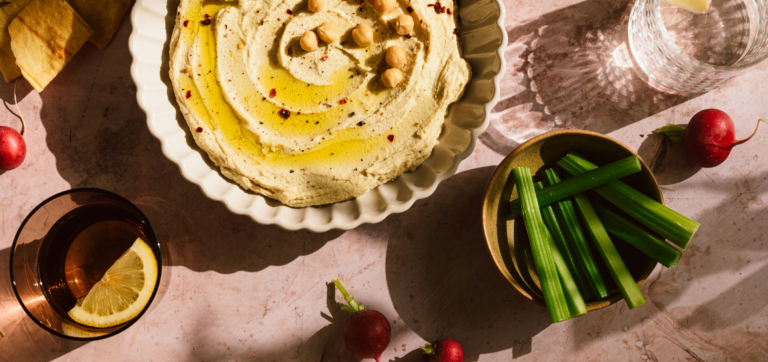 Bowl of hummus with olive oil and chickpeas, celery sticks, radishes, lemon water, and pita chips.