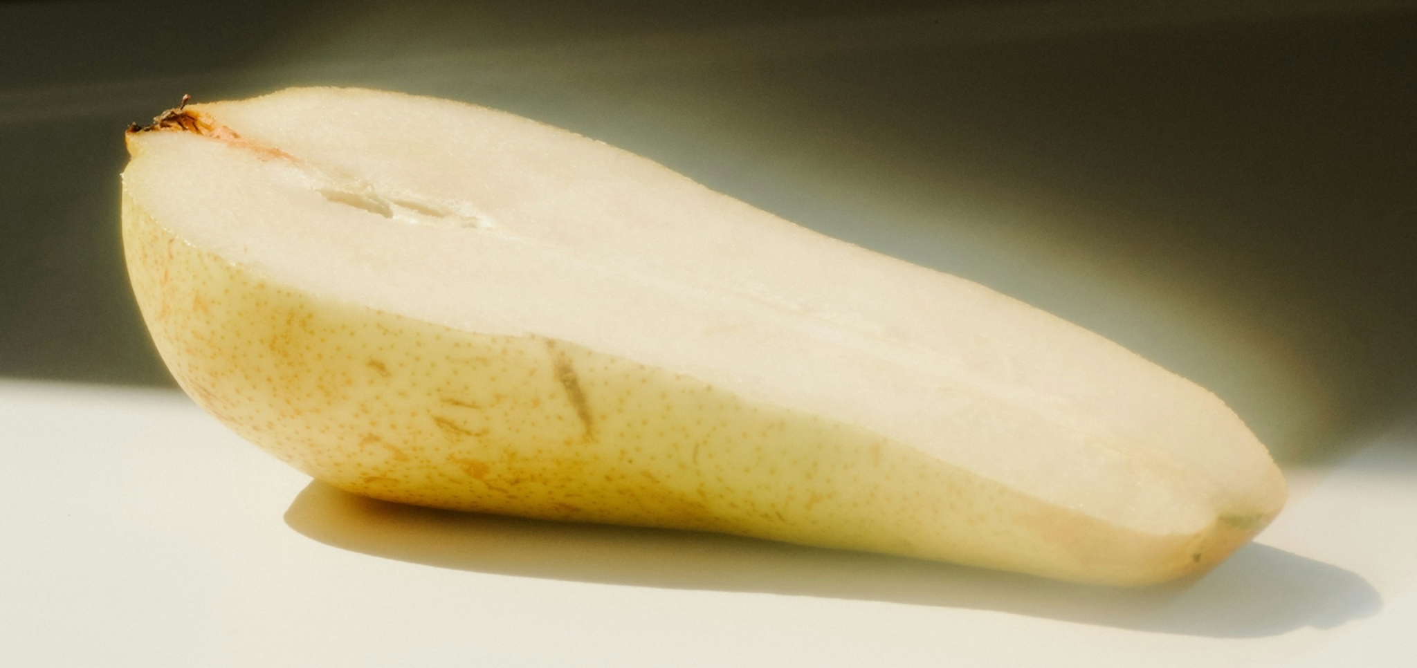 Half of a yellow pear with white flesh on a white surface and dark background.
