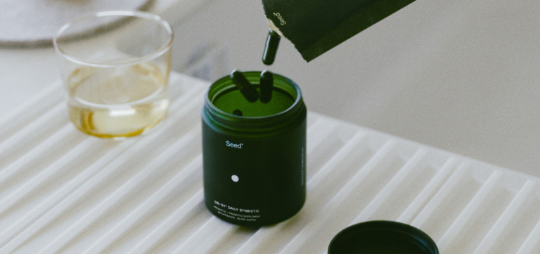 Green supplement bottle with capsules being poured in, next to a glass of liquid.